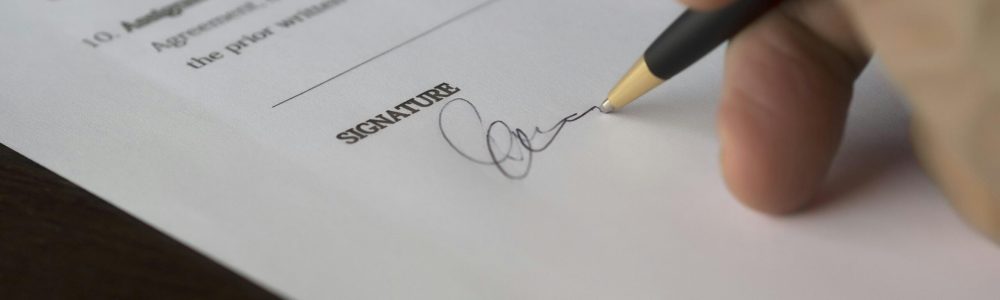 A hand signs a formal contract with a pen on a wooden desk.